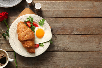 Plate with tasty fried egg, croissant, cup of coffee and rose flower on wooden background