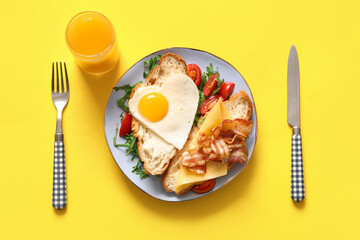 Plate with tasty fried egg, cut croissant, bacon, tomatoes and glass of juice on yellow background