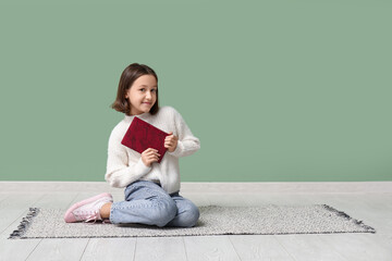 Little girl reading books while sitting on floor near green wall
