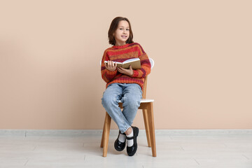Little girl reading book while sitting on chair near beige wall