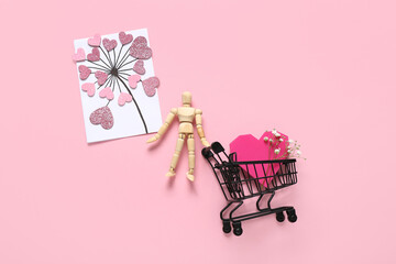Small shopping cart with wooden human figurine, festive postcard and decor on pink background. Valentine's Day celebration