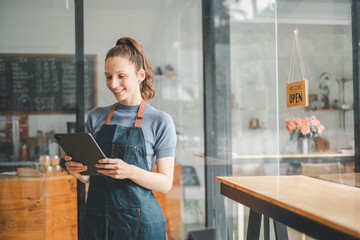 Beautiful young barista woman in apron holding tabltet and standing in front of the door of cafe with open sign board. Business owner startup concept.