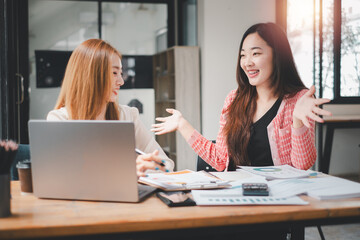 Cheerful businesspeople using a laptop in an office. Happy young entrepreneurs smiling while working together in a modern workspace. Two young businesspeople sitting together at a table.