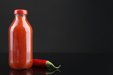 Spicy chili sauce in bottle and pepper against dark background, space for text