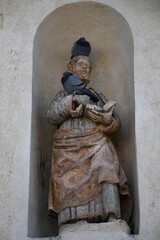 A coloured stone statue of St Peter is in an alcove in a wall. He holds an open book in one hand and a set of keys in another. A pigeon sits on his head, and another on his hand.