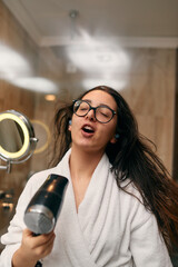 A joyful woman, with headphones in her ears, playfully dries her hair with a hairdryer in the bathroom after a refreshing bath, embodying a carefree and energetic moment of self-care and relaxation