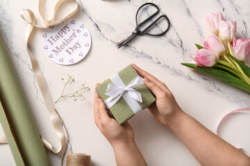 Female hands with gift box, greeting card, scissors and tulip flowers for Mother's Day celebration on light background