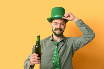 Young man in leprechaun hat with green beard holding bottle of beer on yellow background. St....