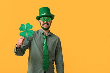Young man in leprechaun hat and decorative glasses with green beard holding clover on yellow...
