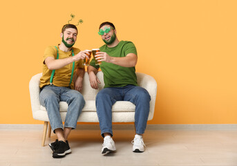 Young men in glasses in shape of clover with green beards holding mugs of beer and sitting on sofa...