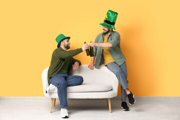 Young men in leprechaun hats with green beards holding bottles of beer and sitting on sofa near...