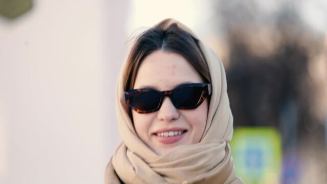 beautiful woman in winter on a sunny day in sunglasses and a warm jacket walks around the city in a headscarf.