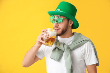 Young man in leprechaun hat and decorative glasses in shape of clover with glass of beer on yellow...