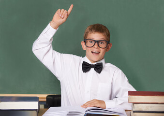 Boy child, books and raised hand in portrait with smile for question, information and knowledge on classroom. Student kid, happy and excited for education, learning and development by chalkboard