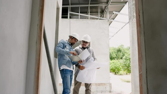 Two Asian men engineers construction supervisors workers  architects designers pair discussed structural drawings notebook  needededited pointed out various points building. Wear white safety helmet.