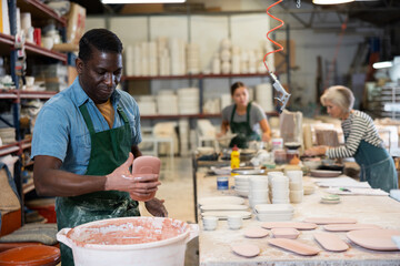 African-american man ceramist painting crafted plates by dipping it into bucket of color.