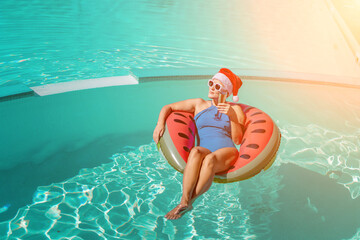 A happy woman in a blue bikini, a red and white Santa hat and sunglasses poses in the pool in an inflatable circle with a watermelon pattern, holding a glass of champagne in her hands. Christmas