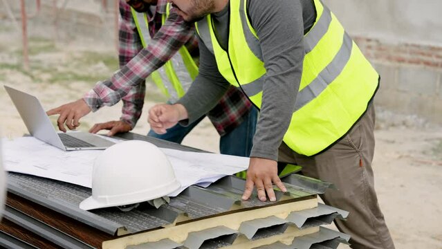 Male workers discuss building plan. Asian engineers in yellow vests work in team comparing plan on laptop and document on table outdoor in front unfinished cement building. They agree on plan