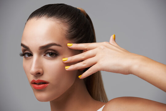 Woman face, beauty and hand with manicure, makeup and yellow nail polish, skin and cosmetics on grey background. Orange lipstick, color nails and model in portrait, confidence in studio and glamour