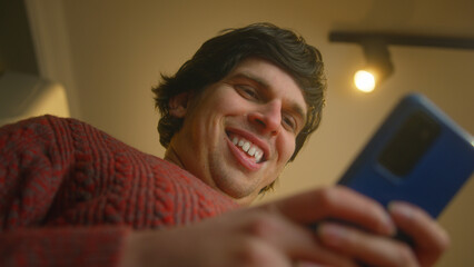 Low angle shot of smiling young man standing in living room typing text message with his...