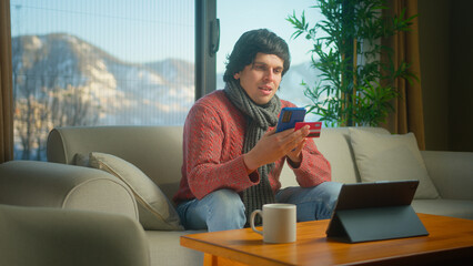 Young man wear scarf holding credit card and smartphone enters credit card number to online...