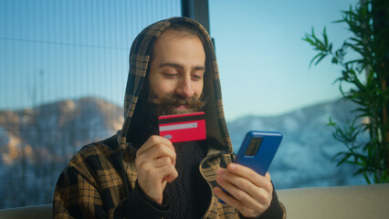 Smiling man in hoodie holding credit card and smartphone enters credit card number to online...