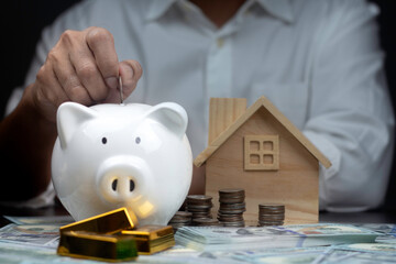 Man putting coin into White piggy bank with wooden house on wood table. saving money for buying house, financial plan home loan concept.