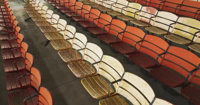 Drone, aerial, video of old faded orange and yellow painted wooden seats at a abandoned a stadium grandstand.
