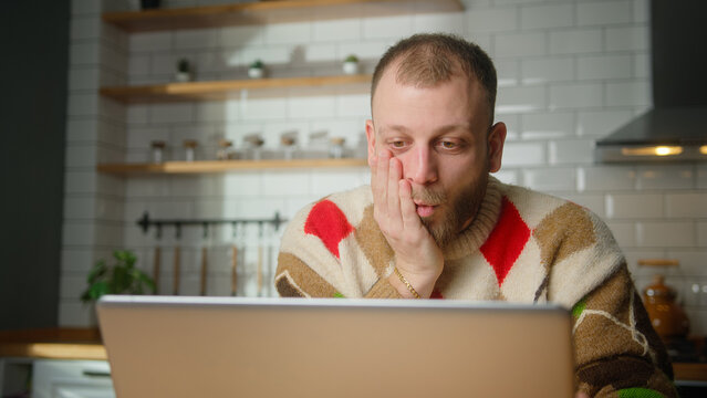 adult man sit in kitchen surprised, stunned while watching movie or video or reading news on laptop at home