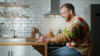 Smiling man sitting in the kitchen having video call conversation through tablet device. Share news, talk to family, enjoy web chat virtual meeting. Side shot