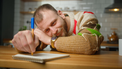 Bored adult man lying on table desk in kitchen drawing in notebook. Man writing on paper, throw...