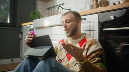 Adult man holding credit card and tablet and due insufficient funds experiencing problems during payment online with credit card while sitting on the floor in kitchen