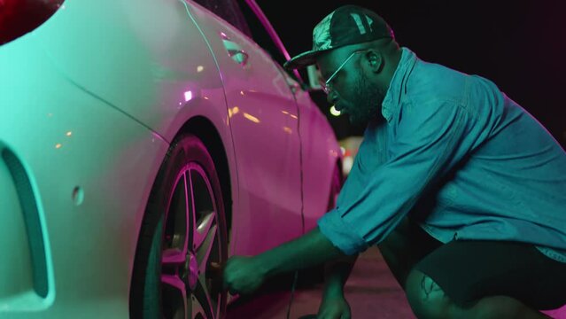 Medium tilt footage of young African American man measuring tyre pressure using air pressure gauge outdoors at night