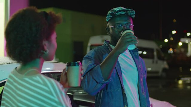 Over shoulder footage of stylish African American guy drinking coffee or tea and talking to unidentified Black lady standing near white car at neon lit fueling station at night