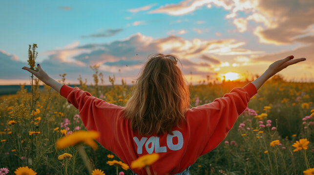 YOLOの生き方を実践している「YOLO」と書かれた赤いトレーナーを着て、お花畑の朝日の太陽に向かって手を広げている女性の後ろ姿