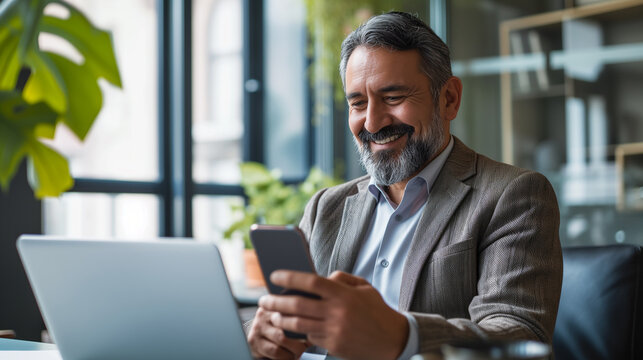 Senior middle aged Hispanic businessman manager ceo using smartphone and laptop in office working online