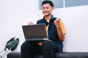 Excited young Asian student with braces looking at laptop on his lap and clenched fist celebrating...