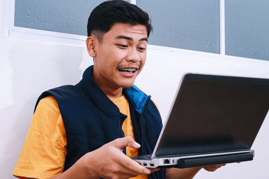 Happy young Asian student man with braces having virtual online learning on laptop computer