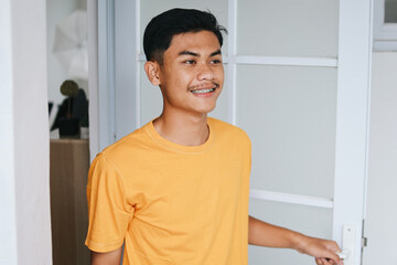Asian young guy with braces open his front door welcoming guest in his house
