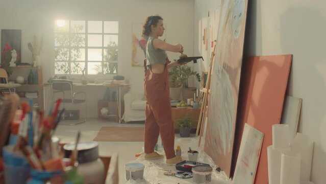 Full side shot of female decorator in jumpsuit standing in studio, holding tray with paint and drawing abstract lines on plywood sheet with wide brush