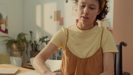 Medium closeup tilting down shot of busy Caucasian woman with disability sitting in wheelchair at table in workshop and drawing abstract lines and shapes in sketchbook with pastels - Powered by Adobe