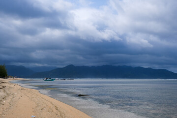 Cloudy sky over a calm beach with boats and a mountain range in the distance.