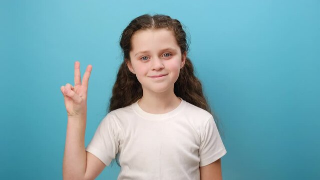Portrait of adorable little girl kid showing v sign with double fingers, gesturing victory, celebrating success, wearing white casual t-shirt, posing isolated over blue color background wall in studio