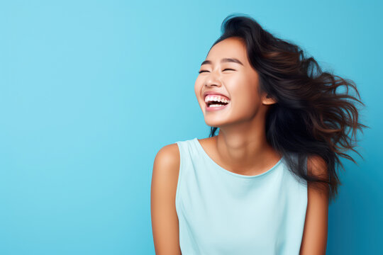 
Photograph of a cheerful young Asian woman laughing heartily, with ample copy space on the left, against a light pastel blue background