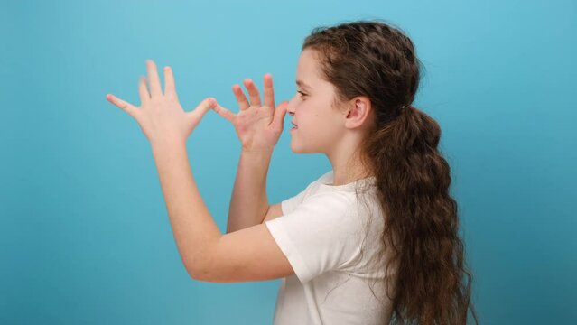Profile portrait of funny caucasian preteen girl child teasing someone, playing with hands near nose, fooling around aside, posing isolated on blue color background wall in studio. Kids humor concept