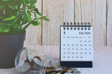 February 2024 desk calendar with stack of coins and potted plant on wooden desk