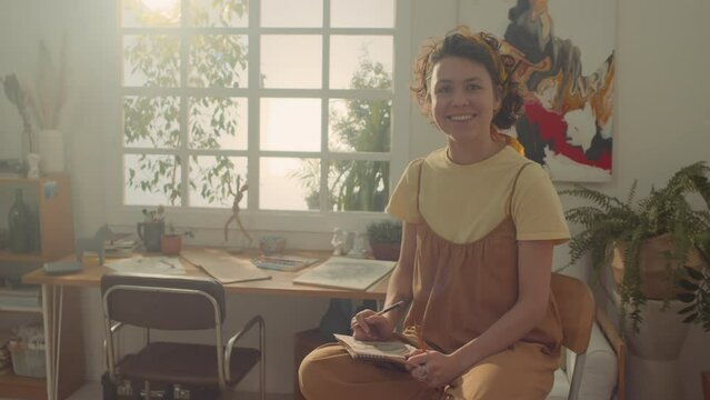 Medium overexposed portrait shot of cheerful young Caucasian female artist with curly hair, in jumpsuit sitting on high chair in workshop, holding sketchbook and pencil, and smiling for camera