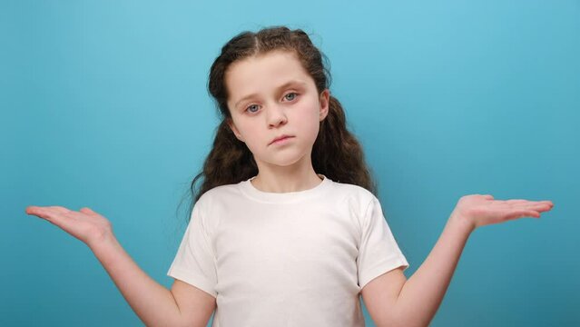 Portrait of unhappy little girl child raising hands in questioning gesture, unsure of answer, expressing doubts, wearing white t-shirt, posing isolated over blue color background wall in studio