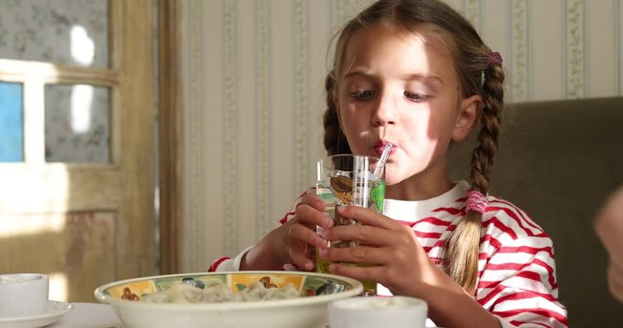 Girl with plaits drinks water from glass with straw before eating sitting at table near mother. Sun rays fall on cheerful child in light premise