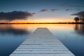 Fototapeta premium Snow-covered jetty in Lake Dirkshorn with a snow shower on the horizon. The layer of snow that fell during the cold night has remained on the jetty.